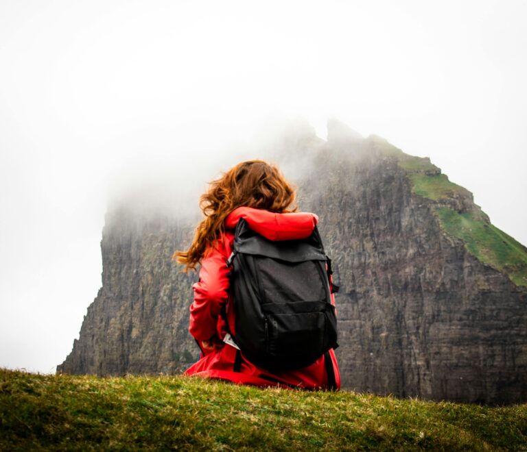A girl sitting in red jacket in front of mountain with a bag
