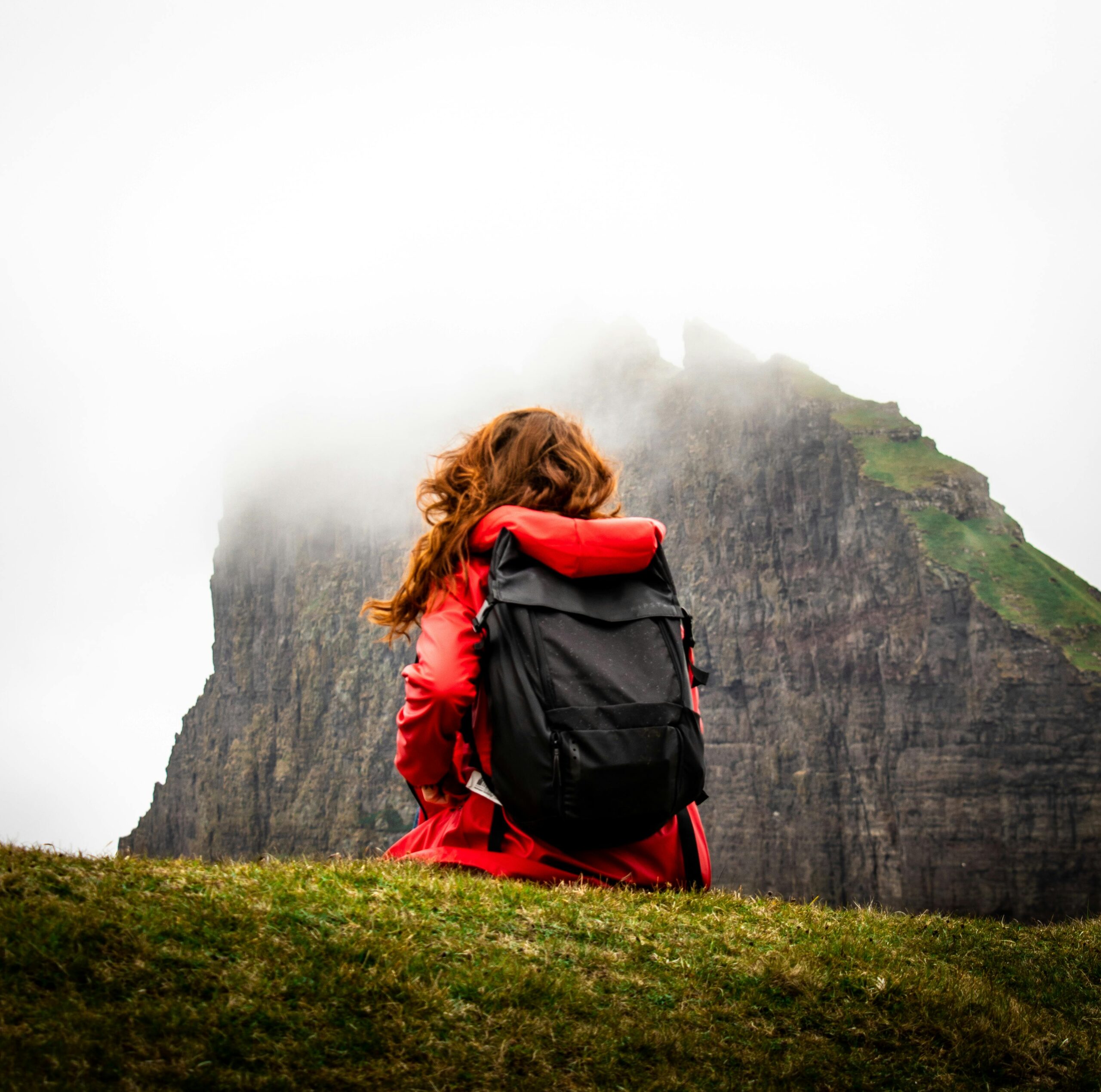 A girl sitting in red jacket in front of mountain with a bag